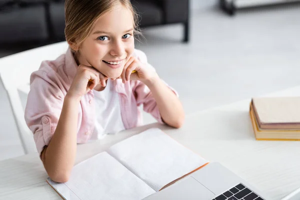 High angle view of cute child smiling at camera near laptop and copy book on table — Stock Photo