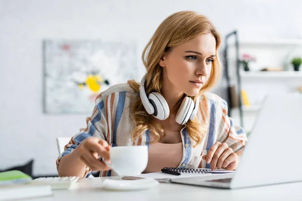 Selective focus of pretty adult freelancer holding cup and working with laptop — Stock Photo