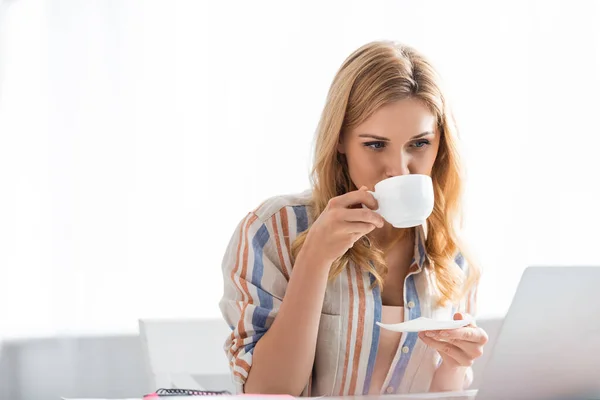 Foco seletivo da mulher bonita bebendo café enquanto trabalhava com laptop — Fotografia de Stock