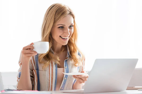 Selective focus of woman smiling to laptop and holding cup — Stock Photo