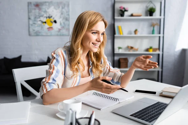 Selective focus of attractive woman smiling and gesturing near laptop — Stock Photo