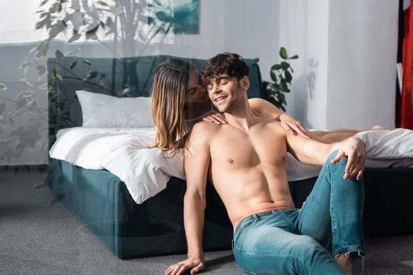 Atractiva mujer besándose sonriente novio sin camisa en el suelo cerca de la cama - foto de stock