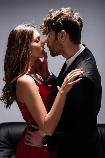 Seitenansicht eines gutaussehenden Mannes im Anzug, der die Lippen einer schönen Frau im roten Kleid in der Nähe von Couch auf grau berührt — Stockfoto