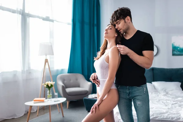 Handsome man undressing sexy woman in bodysuit in bedroom — Stock Photo
