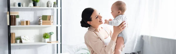 Panoramaaufnahme einer fröhlichen Mutter, die ihren süßen Säugling im Babystrampler in den Armen hält — Stockfoto