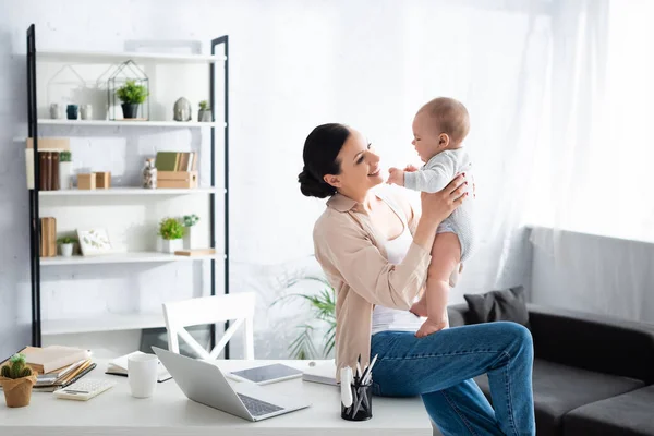 Mãe feliz segurando nos braços filho bebê bonito perto de gadgets e xícara de mesa — Fotografia de Stock