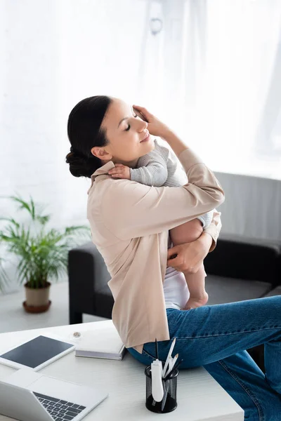 Attractive mother holding in arms and hugging cute infant son near gadgets — Stock Photo