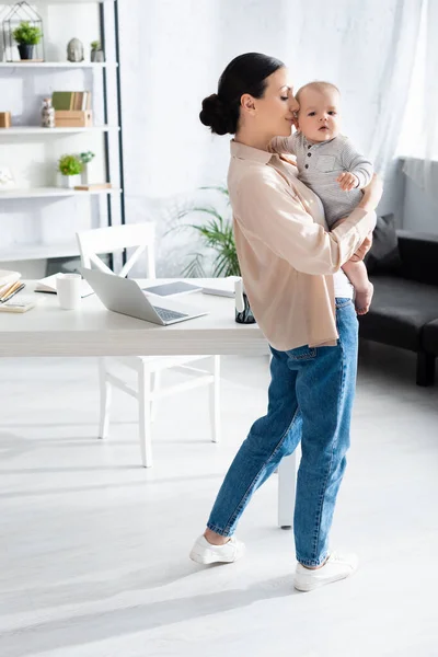 Attraente madre in piedi e tenendo in braccio carino figlio neonato in pagliaccetto vicino gadget sul tavolo — Foto stock