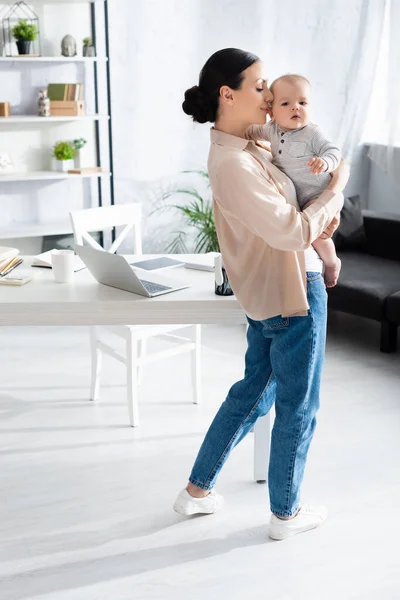 Atraente mãe de pé e segurando nos braços bonito bebê filho no bebê romper perto de gadgets na mesa — Fotografia de Stock