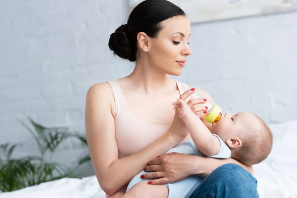 Schöne Mutter füttert niedlichen Säugling Sohn, während sie Babyflasche hält — Stockfoto