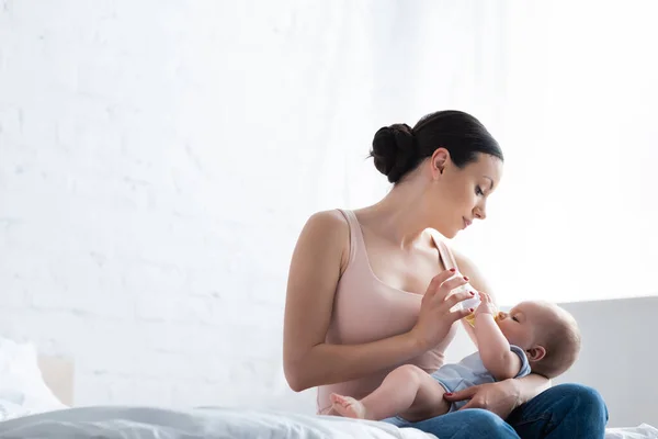 Beautiful woman feeding cute infant son while holding baby bottle — Stock Photo