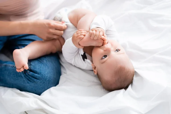 Foco seletivo da mãe vestindo bebê romper em bebê filho chupando pés descalços — Fotografia de Stock