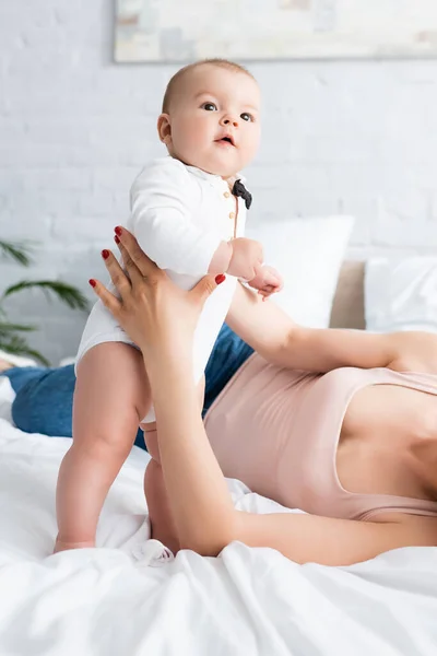 Mother holding cute infant son in baby romper standing on bed — Stock Photo