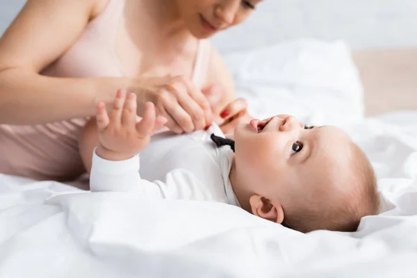 Foyer sélectif de mère touchant le fils de bébé dans la barboteuse de bébé avec noeud papillon sur le lit — Photo de stock