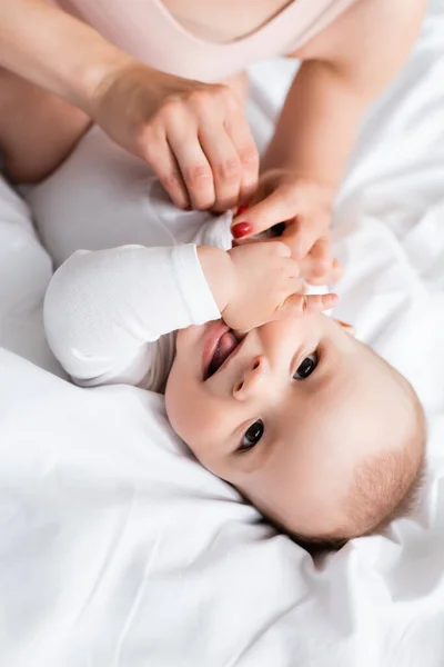 Vista aérea de la madre tocando feliz bebé hijo en bebé mameluco en la cama - foto de stock