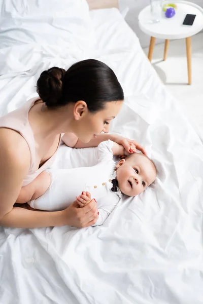 Vista aérea de la madre tocando adorable hijo en bebé mameluco con pajarita acostado en la cama - foto de stock