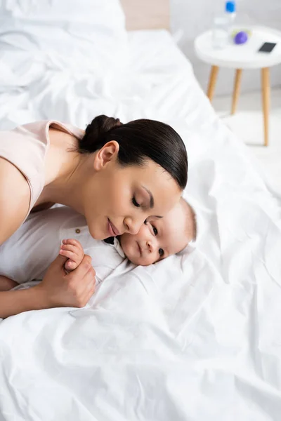 Atractiva madre con los ojos cerrados abrazando adorable hijo en bebé mameluco acostado en la cama - foto de stock