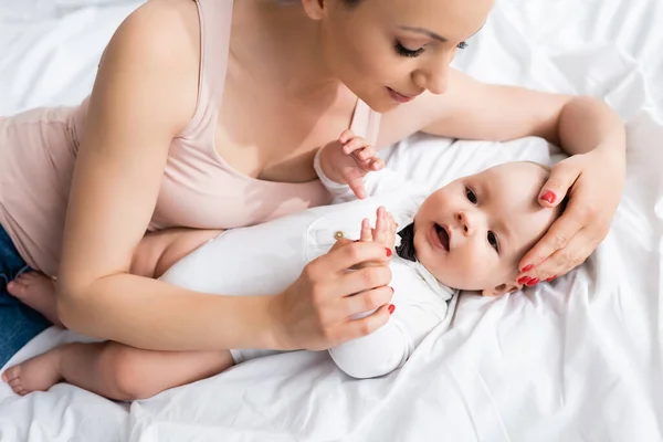 Overhead view of attractive mother touching adorable infant son in baby romper lying on bed — Stock Photo