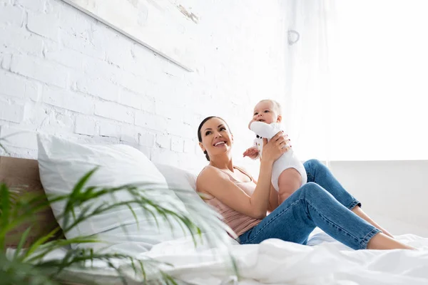 Selektiver Fokus der glücklichen Mutter in Jeans, die auf dem Bett sitzt und den niedlichen Säugling im Arm hält — Stockfoto