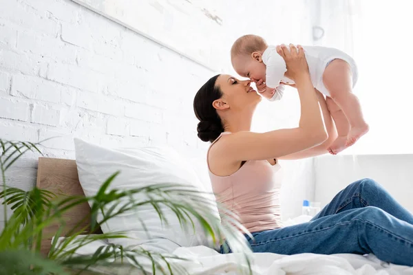 Selektiver Fokus der fröhlichen Mutter in Jeans, die auf dem Bett sitzt und den niedlichen Säugling im Arm hält — Stockfoto