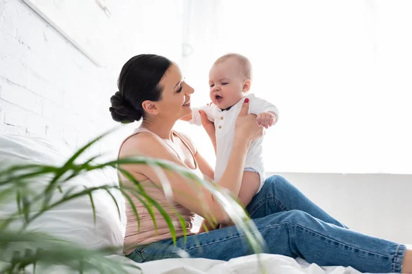 Selective focus of happy woman in jeans sitting on bed and holding in arms cute infant son — Stock Photo