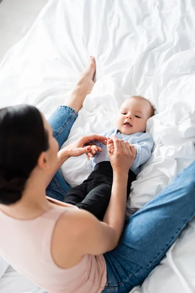 Overhead view of mother in denim jeans sitting on bed and touching cute infant son — Stock Photo