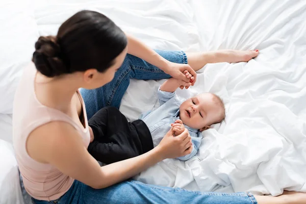 Ansicht der Mutter in Jeans, die auf dem Bett sitzt und die Hände ihres kleinen Sohnes berührt — Stockfoto