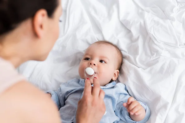Top view of baby boy with pacifier lying on bed and looking at mother — Stock Photo
