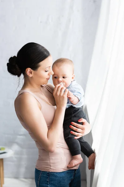 Красива мати тримає в руках босоніж немовля сина в дитячому одязі — стокове фото