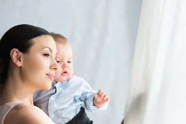 Bela mãe segurando nos braços bonito bebê filho em roupas de bebê e olhando para longe — Fotografia de Stock