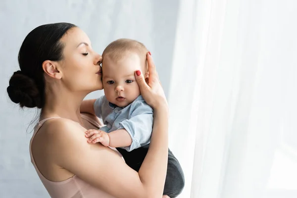 Mãe segurando nos braços e beijando o filho bebê bonito em roupas de bebê — Fotografia de Stock