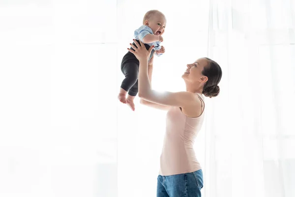Fröhliche Mutter, die sich in den Armen hält und den niedlichen Säugling in Babykleidung betrachtet — Stockfoto