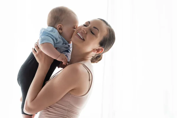 Madre sorridente con gli occhi chiusi tenendo in braccio carino figlio neonato in abbigliamento per bambini — Foto stock