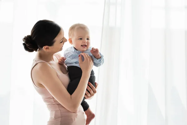 Sorridente mãe segurando nos braços bonito bebê filho em roupas de bebê — Fotografia de Stock