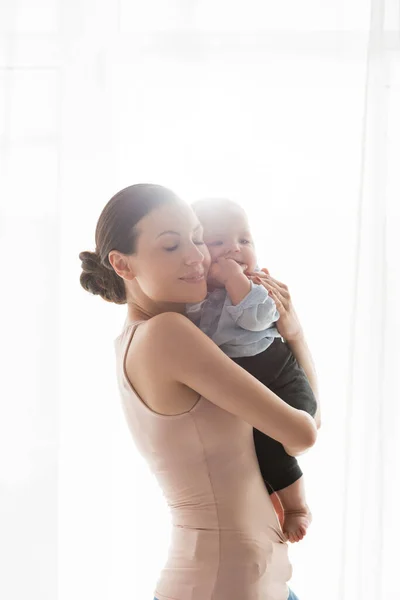 Sonriente madre con los ojos cerrados sosteniendo en brazos adorable infante hijo en ropa de bebé - foto de stock