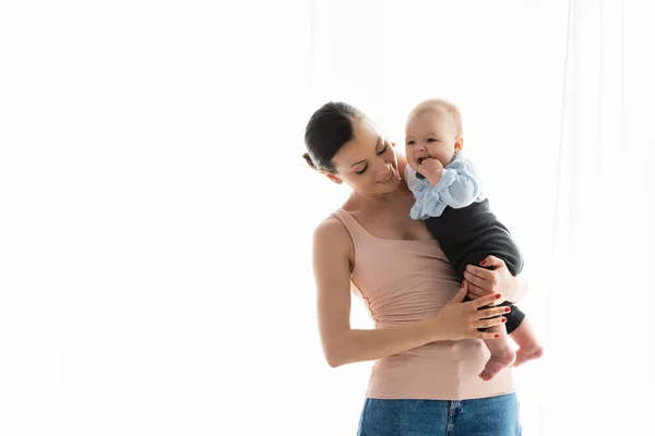 Souriant mère tenant dans les bras pieds nus bébé fils dans les vêtements de bébé — Photo de stock