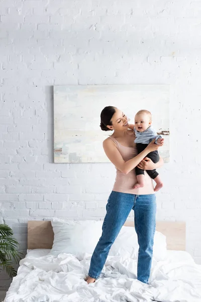 Happy mother holding in arms happy barefoot infant son in baby clothing and standing on bed — Stock Photo