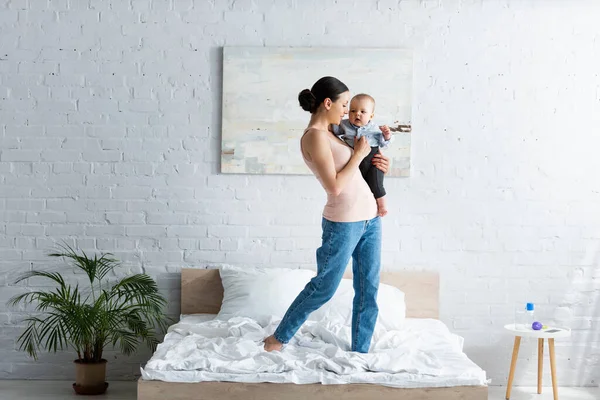 Smiling mother holding in arms barefoot infant son in baby clothing and standing on bed — Stock Photo