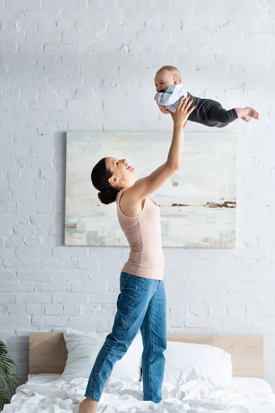 Smiling mother holding barefoot infant son above head and standing on bed — Stock Photo