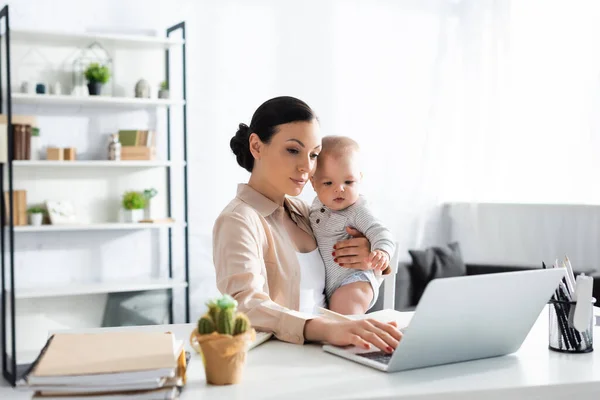 Enfoque selectivo de madre atractiva sosteniendo en brazos bebé niño y trabajando desde casa - foto de stock