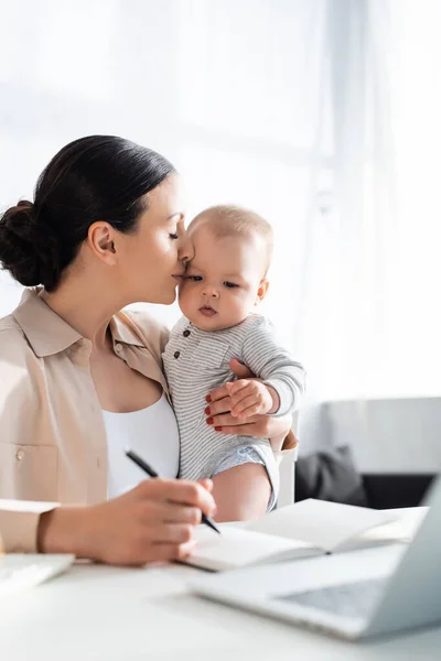 Messa a fuoco selettiva della madre attraente che tiene in braccio e bacia il bambino vicino al laptop — Foto stock