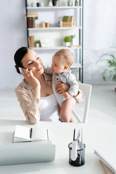 Избирательный фокус привлекательного фрилансера, держащего в руках младенца сына и разговаривающего на смартфоне — стоковое фото