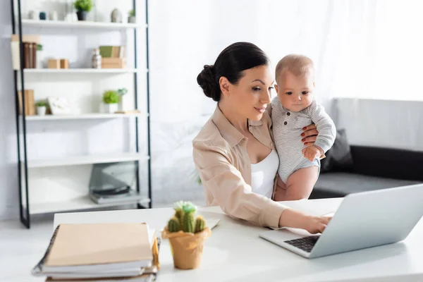 Selektiver Fokus der attraktiven Mutter, die den kleinen Jungen im Arm hält und Laptop benutzt — Stockfoto