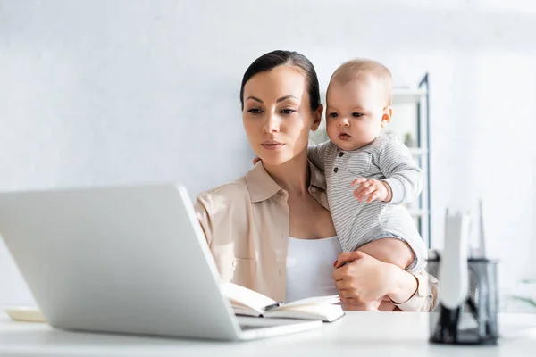 Selective focus of freelancer holding in arms infant son and looking at laptop — Stock Photo