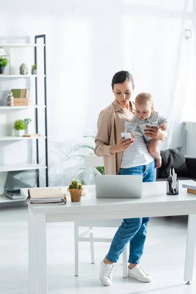 Freelancer sosteniendo en brazos al niño y usando smartphone cerca de laptop - foto de stock