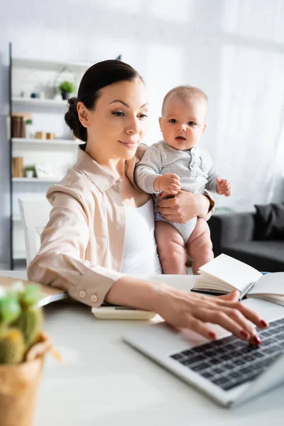 Selective focus of mother typing on laptop keyboard and holding in arms baby boy — Stock Photo