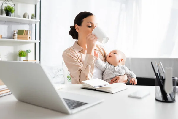 Selective focus of freelancer drinking near cute infant son and gadgets — Stock Photo