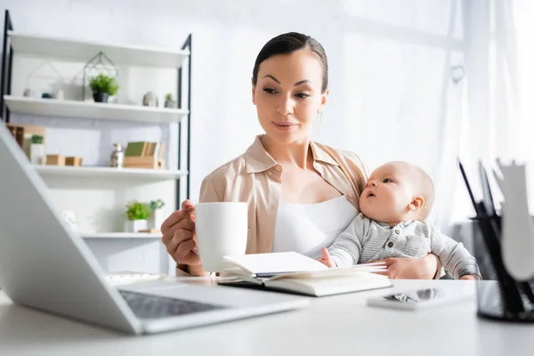 Selective focus of mother holding cup near cute infant son while looking at laptop — Stock Photo