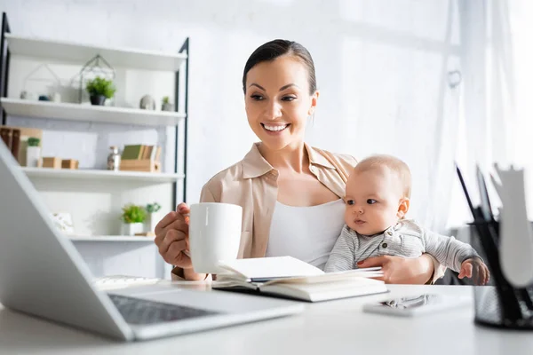 Selective focus of happy mother holding cup near cute infant son and gadgets — Stock Photo