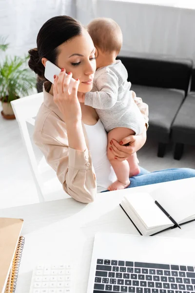 Atractiva madre sosteniendo en brazos hijo lactante y hablando en el teléfono inteligente cerca de la computadora portátil - foto de stock